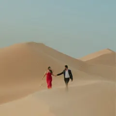 A classic romantic scene of a couple walking hand-in-hand across the crest of a sand dune in Dubai. The bride's flowing red dress provides a beautiful splash of color against the neutral, sweeping landscape.