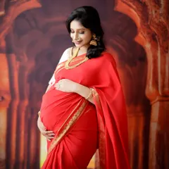 A graceful solo portrait of a mom-to-be in a stunning red saree. Her serene expression and the traditional temple backdrop create a timeless and beautiful image.
