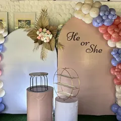 A "He or She" gender reveal party setup. The decor features two arched backdrops and a beautiful balloon garland in a mix of pink, blue, and neutral tones.
