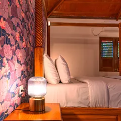 A closer look at the details. The intricate carving on the wooden bedpost, the warm glow of the bedside lamp, and the vibrant floral wallpaper come together to create a tranquil and inviting atmosphere.