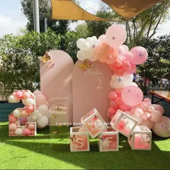 A delicate butterfly-themed birthday decoration in soft pink tones. This outdoor setup includes custom arched backdrops, balloon-filled transparent boxes spelling out the name, and a balloon-filled marquee number.