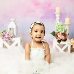 A happy baby girl surrounded by soft clouds and flowers in a whimsical studio setup.