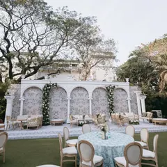 An elegant outdoor lounge area with a European feel, featuring a white arched backdrop with delicate floral patterns, comfortable seating, and classic decor elements.