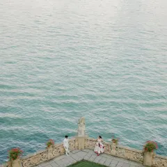 A breathtaking aerial view of a couple on a stone balcony overlooking Italy's Lake Como. The vast, serene turquoise water creates a sense of scale and tranquility, making for an epic and beautiful destination pre-wedding shot.