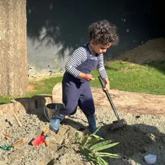 A child dressed as a gardener tends to the sandpit. Our outdoor spaces, including our sand and mud kitchens, are rich environments for imaginative play and connecting with natural elements.