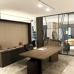 This image captures an office during the final stages of furnishing. A large, minimalist wooden desk is in place, with custom dark wood cabinetry along the wall. The glass partition with a geometric black frame separates the space while maintaining an open feel.