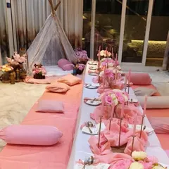 A cute and cozy sit down dinner setup, all in shades of pink. This low seating arrangement features pink floor coverings and cushions, a long table with pink floral centerpieces, and a lace teepee tent in the corner for a whimsical touch.