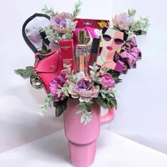 A fun and trendy gift arranged inside a large pink tumbler. This creative bouquet includes lip balm, perfume, an eye mask, a mini pouch, and other small accessories, all held together with faux flowers.