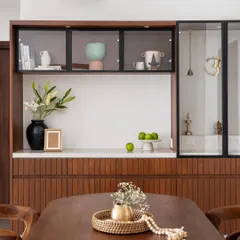 This custom unit serves as both a crockery cabinet and a puja space. The design features fluted wood paneling, a marble countertop, and glass-fronted upper cabinets.