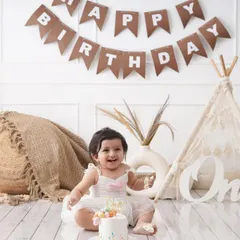 A happy baby girl enjoys her first birthday cake in a boho-themed setup, complete with a miniature tent and a 'One' sign.