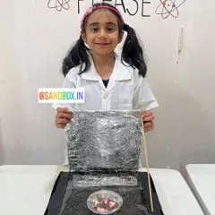 This young scientist is ready to cook with the sun! She carefully constructed her solar cooker using a box, aluminum foil, and plastic wrap to trap the sun's heat.