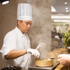 A chef carefully tends to steaming bamboo baskets at a live dim sum station. This showcases our commitment to serving fresh, authentic, and skillfully prepared global cuisine with professional care.