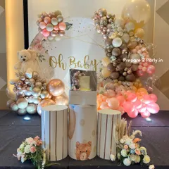 An elegant and modern "Oh Baby" themed baby shower backdrop. The setup features a sophisticated palette of rose gold, pink, and beige balloons, a plush teddy bear, custom pedestals for the cake, and delicate floral arrangements.