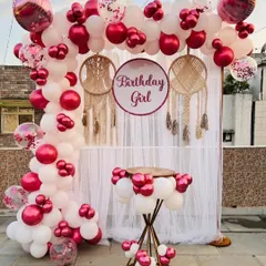 A stylish birthday photo booth on a terrace, with a white curtain backdrop, macrame details, and a vibrant pink and white balloon arch.
