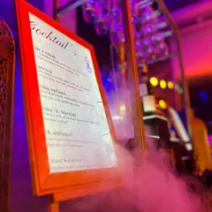 The custom cocktail menu for a Sufi-themed event, featuring poetic drink names like "Ishq Sufiana" and "Mast Kalandar." The menu is presented on an easel amidst atmospheric smoke.