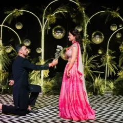 A romantic moment from a Sangeet night. The couple is on a stage with a unique black and white patterned floor and a backdrop of greenery, disco balls, and warm lights.