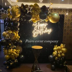 A glamorous black and gold birthday photo booth, featuring a black sequin wall, an elaborate balloon garland, and a neon sign.