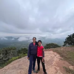A mother and son duo smiling for a photo at the Kabbaldurga summit on a cloudy day. We welcome adventurers of all ages to create special memories together.