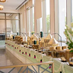 A full view of a luxurious buffet line featuring gleaming golden chafing dishes and decorative urns. The table is adorned with traditional flower garlands, adding a festive touch to this puja celebration.