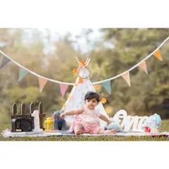 A beautiful setup for baby Anwita's first birthday photoshoot. The scene is complete with a teepee, bunting, a wooden fence, and a "One" sign, all arranged in a lovely park setting.