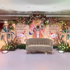 A beautiful stage designed for a religious ceremony, featuring traditional Pichwai art of Shrinathji and his cows. The decor is complemented by floral arrangements and banana leaf motifs, creating a serene and spiritual ambiance.