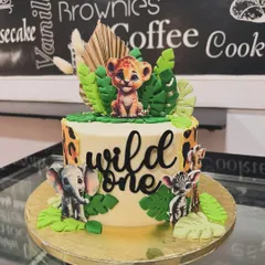 A "Wild One" first birthday cake featuring a baby lion cub, elephant, and zebra. The design incorporates leopard print details and lush green fondant leaves for a complete safari look.