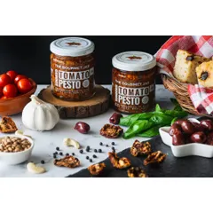 Sundried Tomato Pesto jars by The Gourmet Jar, surrounded by ingredients like tomatoes, olives, and focaccia bread. This composition helps customers imagine how they can use the product.