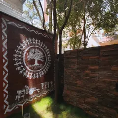 My Tree of Life mural on a compound wall, seen in the daylight. The sunlight reveals the texture of the wall and the clean, white pigment of the Warli painting, which includes a bullock cart scene below the central motif.