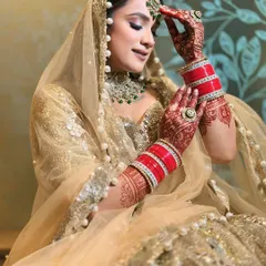 A beautiful candid moment. The bride adjusts her maang tikka, showing off the intricate henna and the way the golden fabric drapes. This lehenga is paired with green emerald jewellery for a classic color combination.