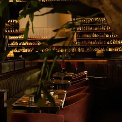 A view of the dining area, framed by the leaves of the olive tree. The glowing, double-height bar in the background serves as a dramatic, amber-lit backdrop.
