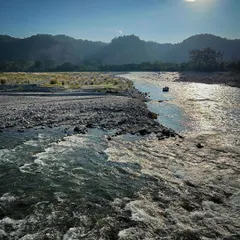 The sun shines brightly over the flowing river and rocky banks in the Himalayan foothills. You can almost feel the warmth of the sun and hear the sound of the water.