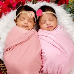 A close-up shot of twin baby girls, swaddled in pink and nestled together among vibrant flowers. Their peaceful expressions show how comfortable they are.