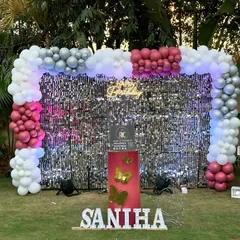 An outdoor party setup on a lawn featuring a silver shimmer wall framed by a balloon arch. The large marquee number '1' and light-up name sign look fantastic as day turns to night.