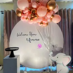 A simple and elegant welcome home backdrop for Ammu. The design features a clean white circular backdrop, a pink and gold balloon arrangement, and a cute elephant cutout.