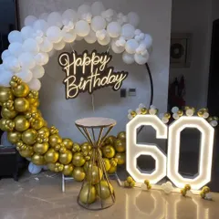 An elegant 60th birthday setup with a gold and white balloon ring, a chic 'Happy Birthday' neon sign, and large light-up numbers.