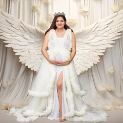 A powerful and angelic studio portrait of an expectant mother wearing a beautiful white gown and a crown. She stands gracefully with large, white angel wings behind her against a dreamy, cloud-like backdrop, portraying the miracle of motherhood.
