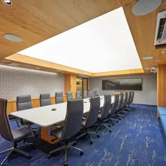A large, formal boardroom featuring a stretch-lit ceiling that provides bright, even, and glare-free light. The room is equipped with a long conference table, comfortable seating, and an additional bench along the wall for larger meetings.