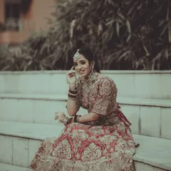 A classic bridal portrait capturing a moment of happy reflection. The bride is seated on outdoor steps, her red and gold lehenga beautifully detailed, showcasing her complete wedding day look.