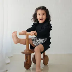 A playful toddler kicks her leg up while riding a wooden tricycle indoors, laughing with excitement.