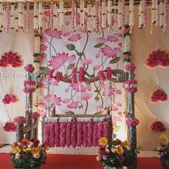 A beautiful naming ceremony design featuring a Pichwai-style lotus painting as the backdrop. The cradle is decorated with pink flowers, and the entire setup is framed by hanging jasmine garlands.