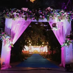 A grand floral entrance arch for a wedding, decorated with purple and pink flowers and delicate hanging bird origami. The dramatic pink lighting and draped fabric create a stunning entryway that leads to the main event.