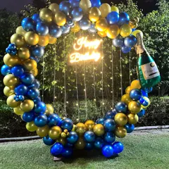 An elegant outdoor birthday celebration setup. The circular frame is decorated with a classy blue and gold balloon garland, complemented by fairy lights and a champagne bottle balloon for a festive touch.