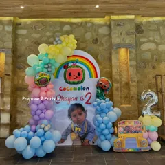 This Cocomelon setup for a second birthday features a custom arch backdrop with a rainbow and personalized photo. The scene is completed with a Cocomelon bus cutout and a pastel balloon garland.