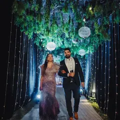 The couple makes a stunning entrance through a magical walkway, with a ceiling of lush greenery and hanging globes, flanked by shimmering curtains of light.