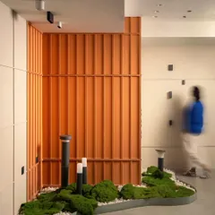 The motion-blurred figure gives a sense of movement and scale to the space. The design combines natural elements like moss with modern industrial materials and bold orange fluted panels.