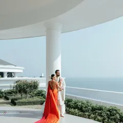 A breathtaking shot of the couple in Vietnam, with the bride's vibrant orange and red saree flowing behind her against the backdrop of the sea and a white villa.