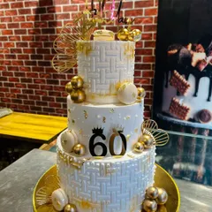 Celebrating a 60th birthday in style with this grand three-tier cake. The design features a white basket-weave texture, gold spheres, macarons, and elegant fan details for a truly luxurious feel.