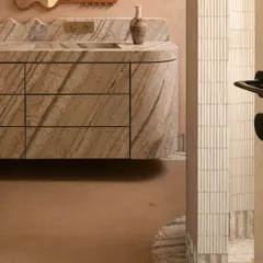 We call this bathroom a 'chic desert dream'. It features blush-toned micro-concrete walls and a monolithic vanity carved from veined Corteccia marble. The stone appears to trickle down onto the floor, creating a fluted skirting detail that is both organic and luxurious.