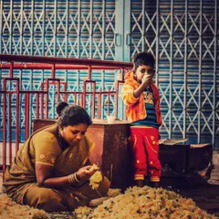 A woman sits on the ground sorting through a pile of flowers, while a small child stands beside her, looking towards the camera. This image tells a story of family and work in a market setting.