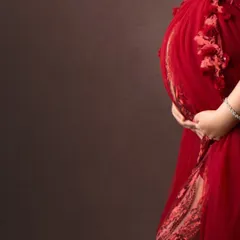 A beautiful close-up detail shot, focusing on the hands cradling the baby bump and the intricate texture of the red gown.